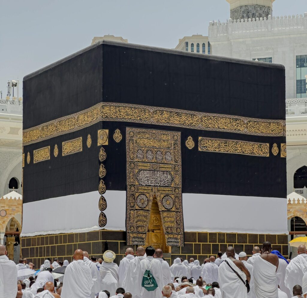Pilgrims in white garments gather around the Kaaba in Mecca during daytime prayers.