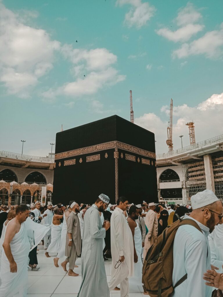 Pilgrims in traditional attire perform Tawaf around the Kaaba in Mecca, Saudi Arabia under a clear blue sky.